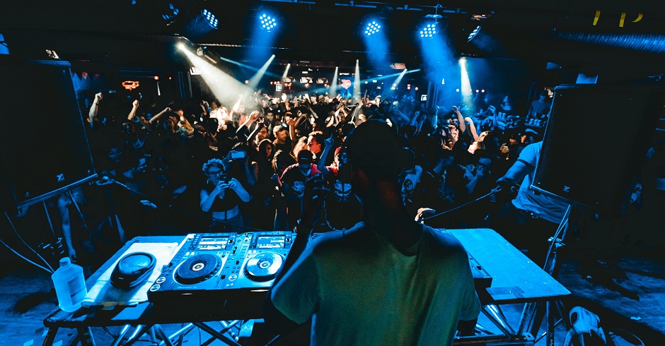 DJ von hinten hinter dem Mischpult. Dunkler Club mit Scheinwerfer, tanzende Menschen.