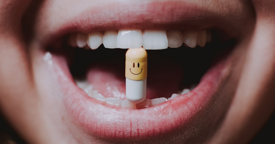 Eine Medikamentenkapsel mit einem Smiley wird von einem lachenden Menschen zwischen den Zähnen gehalten.