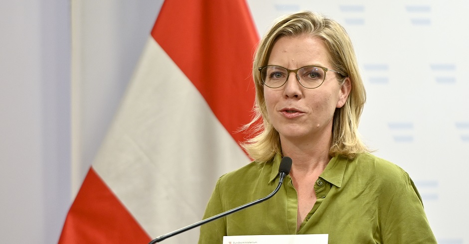 Leonore Gewessler bei einer Pressekonferenz. Sie steht vor einer Österreichfahne und spricht in ein Mikrofon.