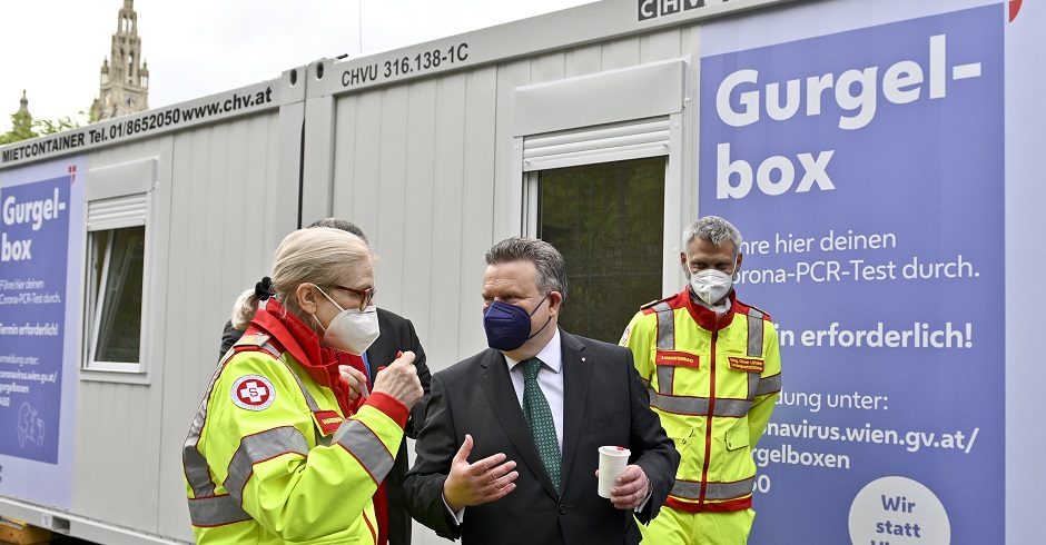 Bürgermeister Michael trägt eine Maske. Er steht mit zwei Sanitätern vor einer Gurgelbox.