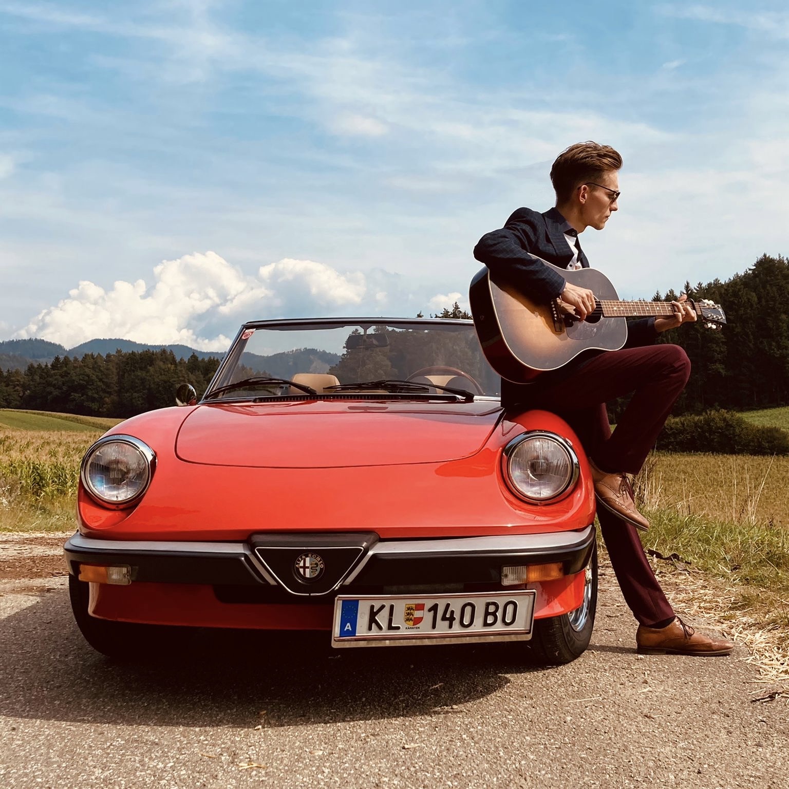Der junge Musiker Simon Stadler lehnt mit einer Akustikgitarre in der Hand an der Motorhaube eines roten Cabrios vor ländlicher Kulisse