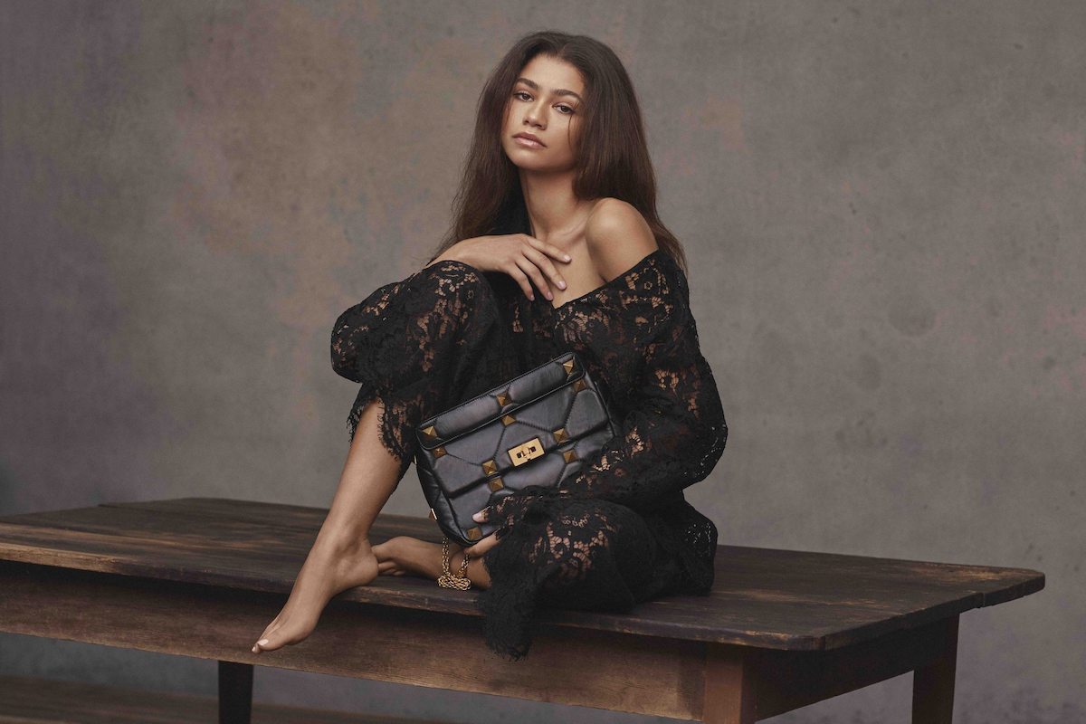 Porträt von Zendaya auf einem Tisch sitzend und Tasche präsentierend