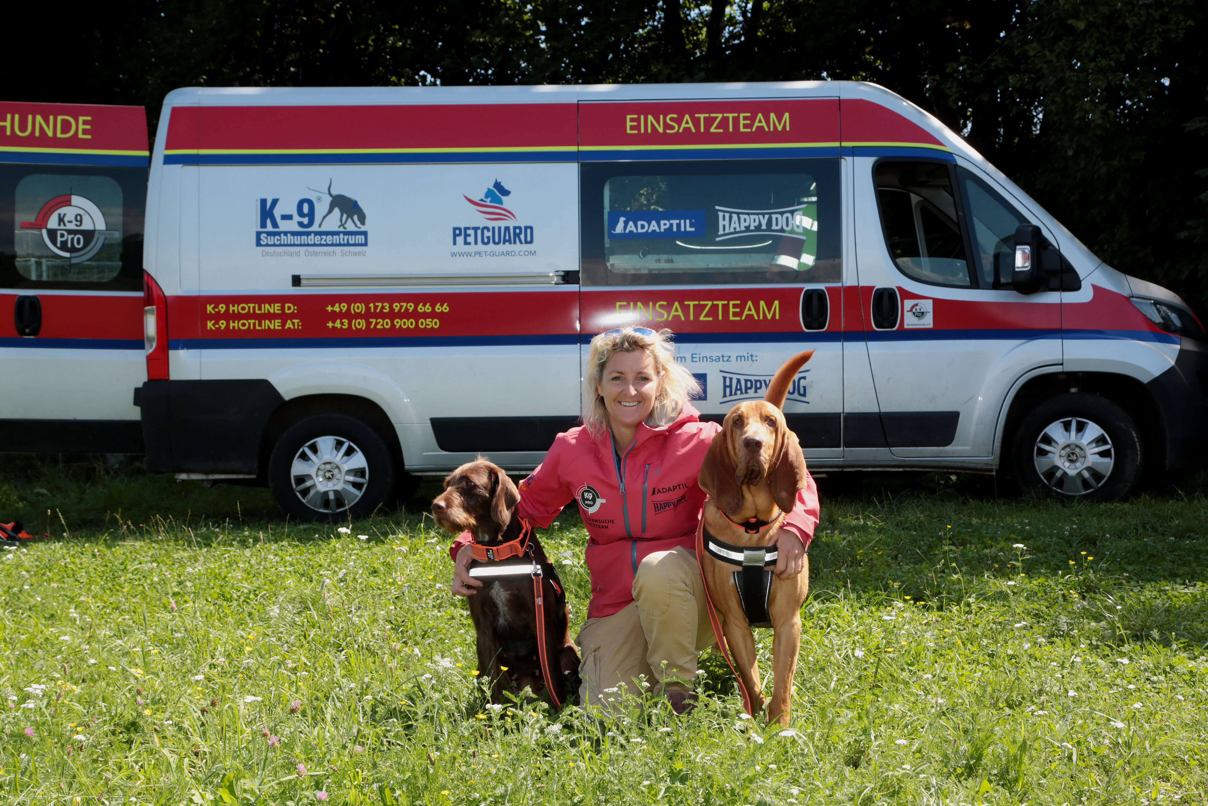 Alexandra Grunow kniet, flankiert von ihren zwei Hunden, auf einer Wiese vor dem Einsatzwagen des K9 Pro Vermisstensuchevereins