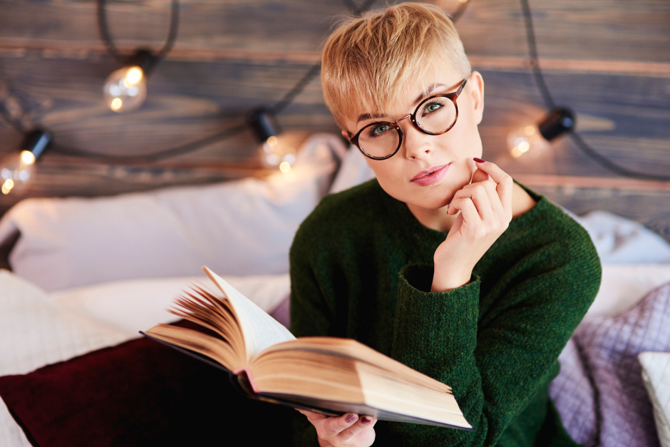 Frau mit Brille und blonder Kurzhaarfrisur blickt von ihrem Buch Richtung Kamera auf | Credit: iStock.com/gpointstudio