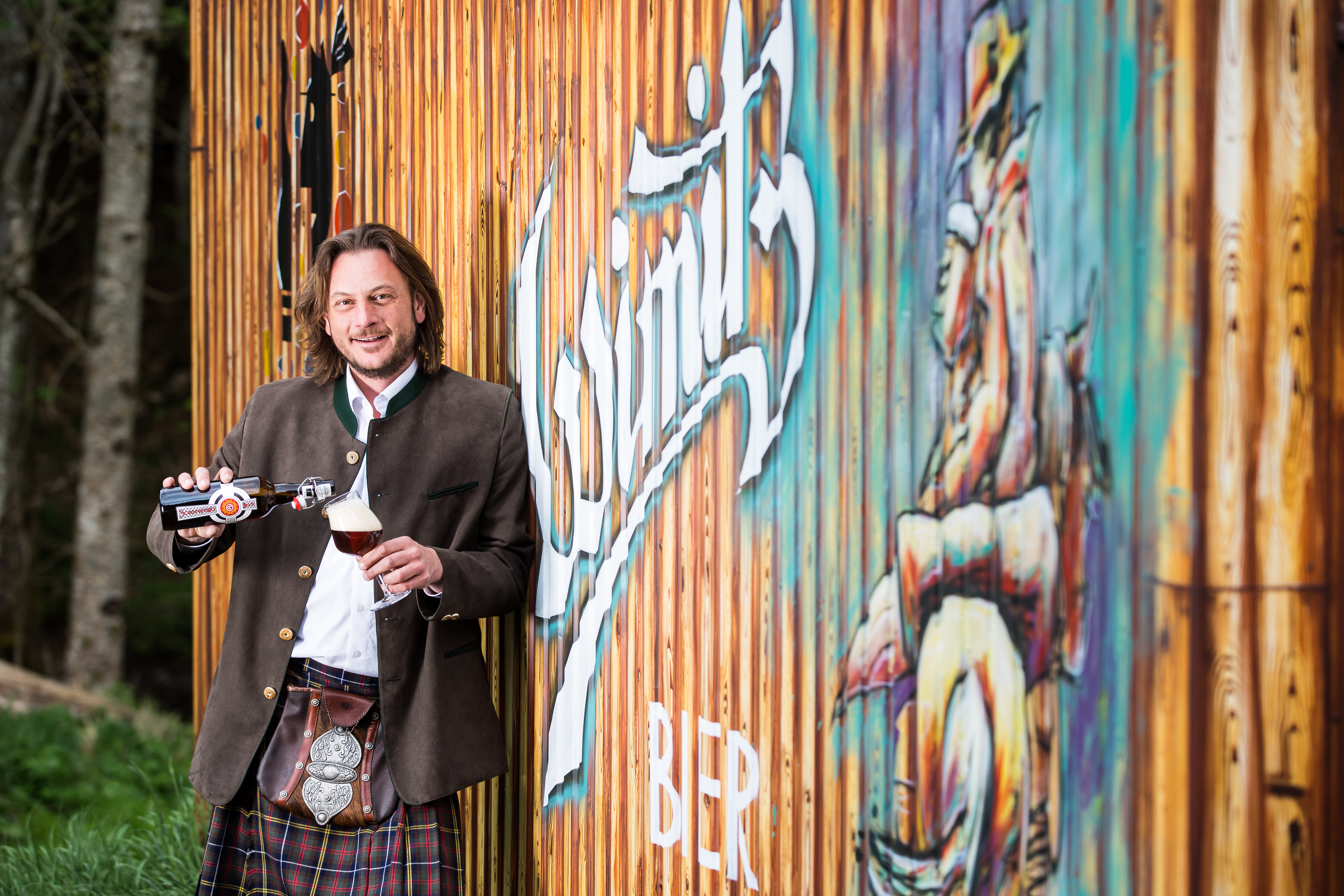Josef Habich steht im Kärntner Kilt vor dem Logo seiner Brauerei und schenkt sich ein dunkles Bier ein