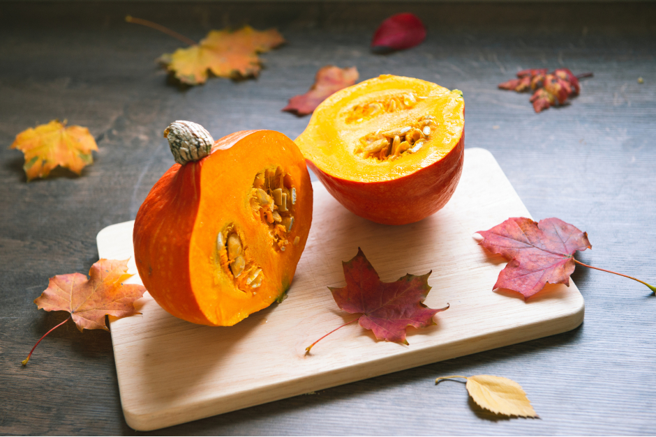 Kürbis in zwei Hälften geteilt auf einem mit herbstlichen Deko-Blättern geschmückten Holzbrett | Credit: iStock.com/petrograd99
