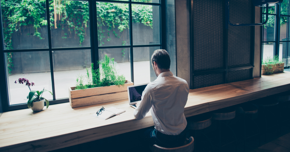 Mann von hinten, der in einem Café vor seinem Notebook sitzt - mit Blick aus dem Fenster auf einen begrünten Innenhof | Credit: iStock.com/Deagreez