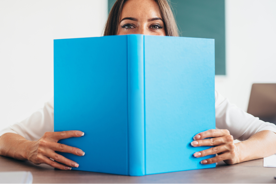 Junge Frau verbirgt ihr Gesicht hinter einem großen Buch mit hellblauem Einband - nur die Augen sind noch zu sehen | Credit: iStock.com/undrey