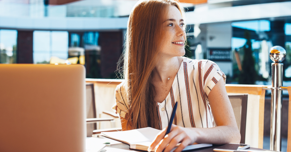 Rothaarige Studentin an einem sonnigen Tag in einem Straßencafé, den Notebook vor sich aufgeklappt | Credit: iStock.coM/Strelciuc Dumitru