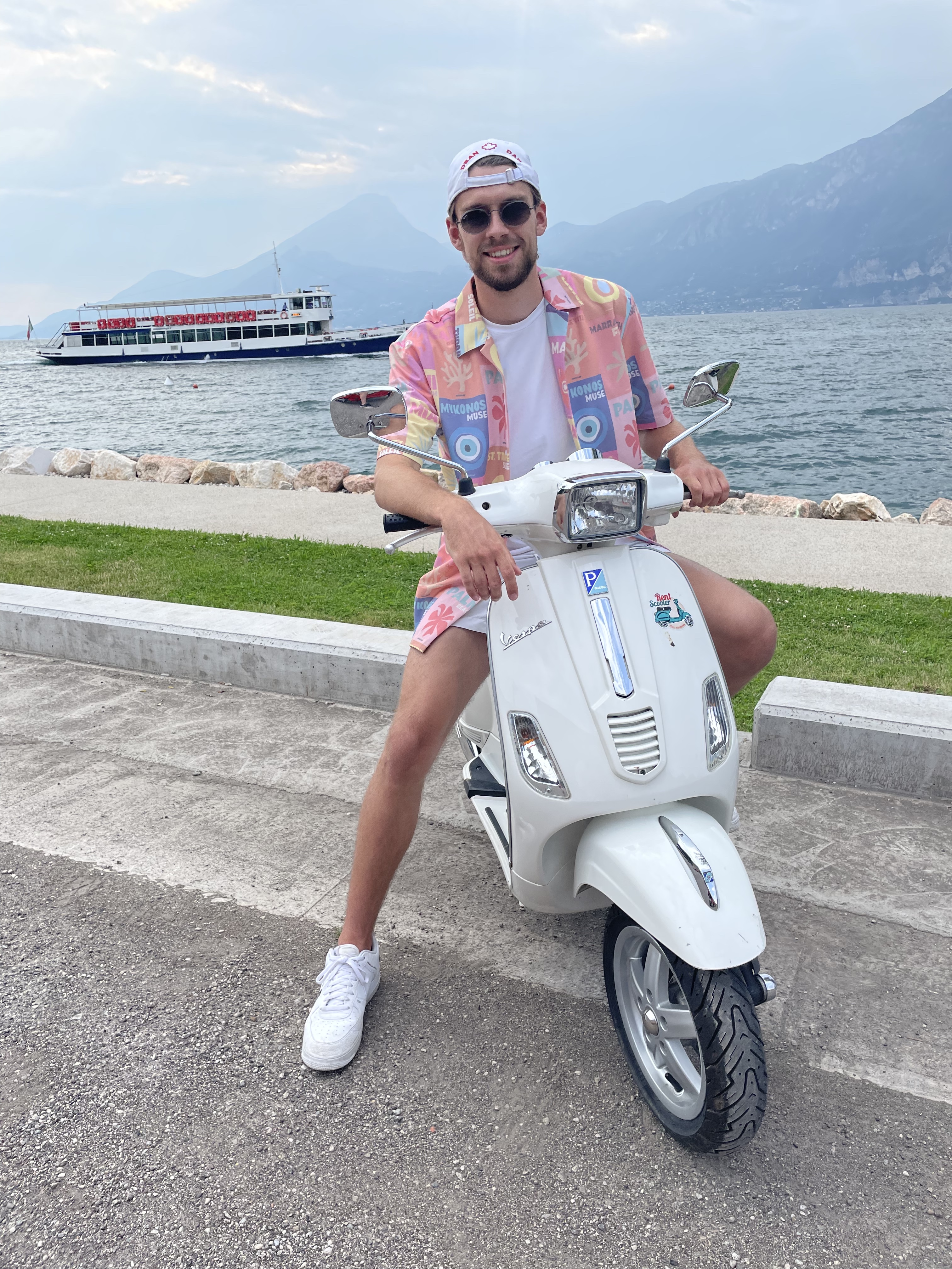 Phillip Menzel in sommerlicher Kleidung auf einer weißen Vespa am Ufer des Wörthersees