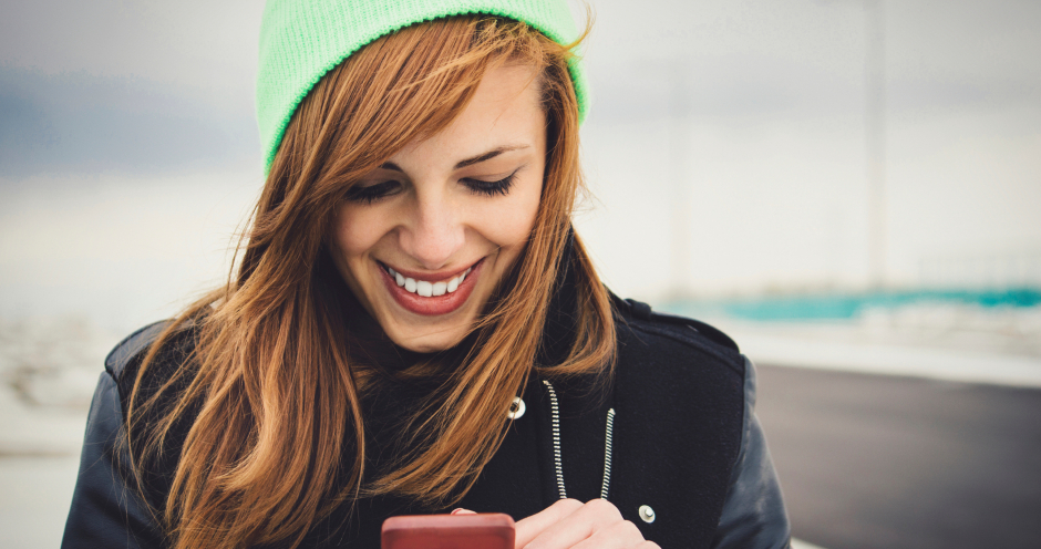 Glückliche Frau blickt während einem Spaziergang an einem kalten Wintertag auf ihr Smartphone | Credit: iStock.com/DjordjeDjurdjevic