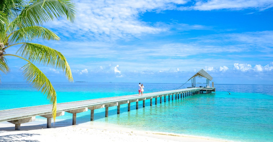Von einem karibischen Sandstrand mit Palme führt ein Steg zu einer kleinen Hütte über dem türkisen Wasser.