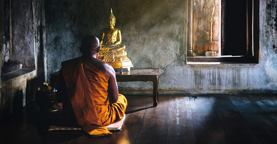 Buddhistischer Mönch in Thailand sitzt im Schneidersitz vor einer Buddha-Statue | Credit: iStock.com/flukyfluky