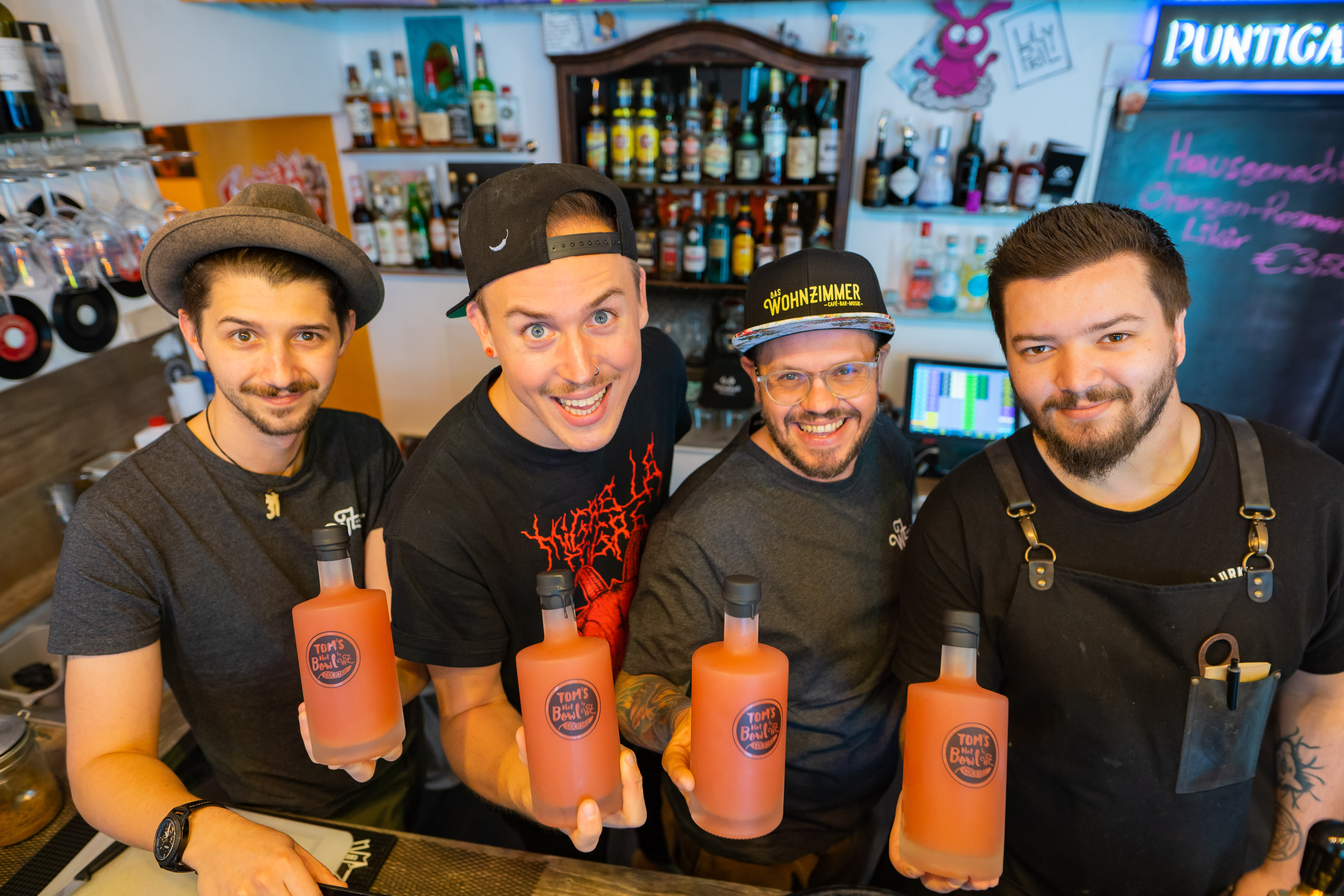 Vier Barkeeper stehen hinter einem Tresen und halten Flaschen mit einem orangen Inhalt in die Kamera