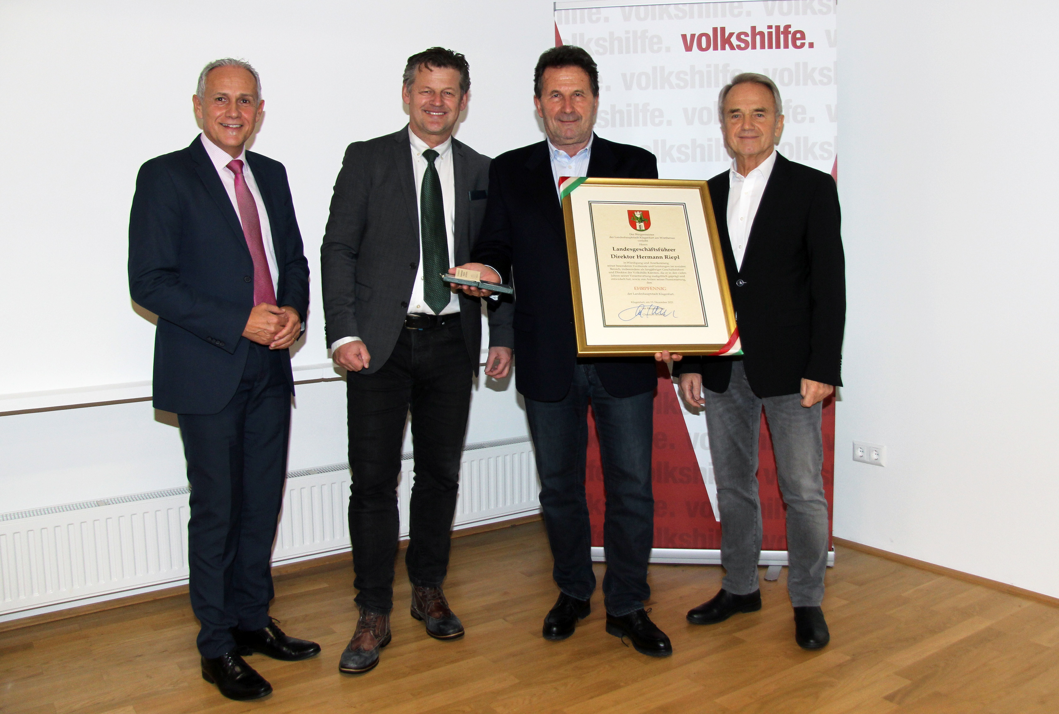 Hermann Riepl steht mit einer Urkunde und dem Ehrpfennig in der Hand neben Bürgermeister Scheider und flankiert von Jürgen Pfeiler und Ewald Wiedenbauer vor einem Rollup der Volkshilfe