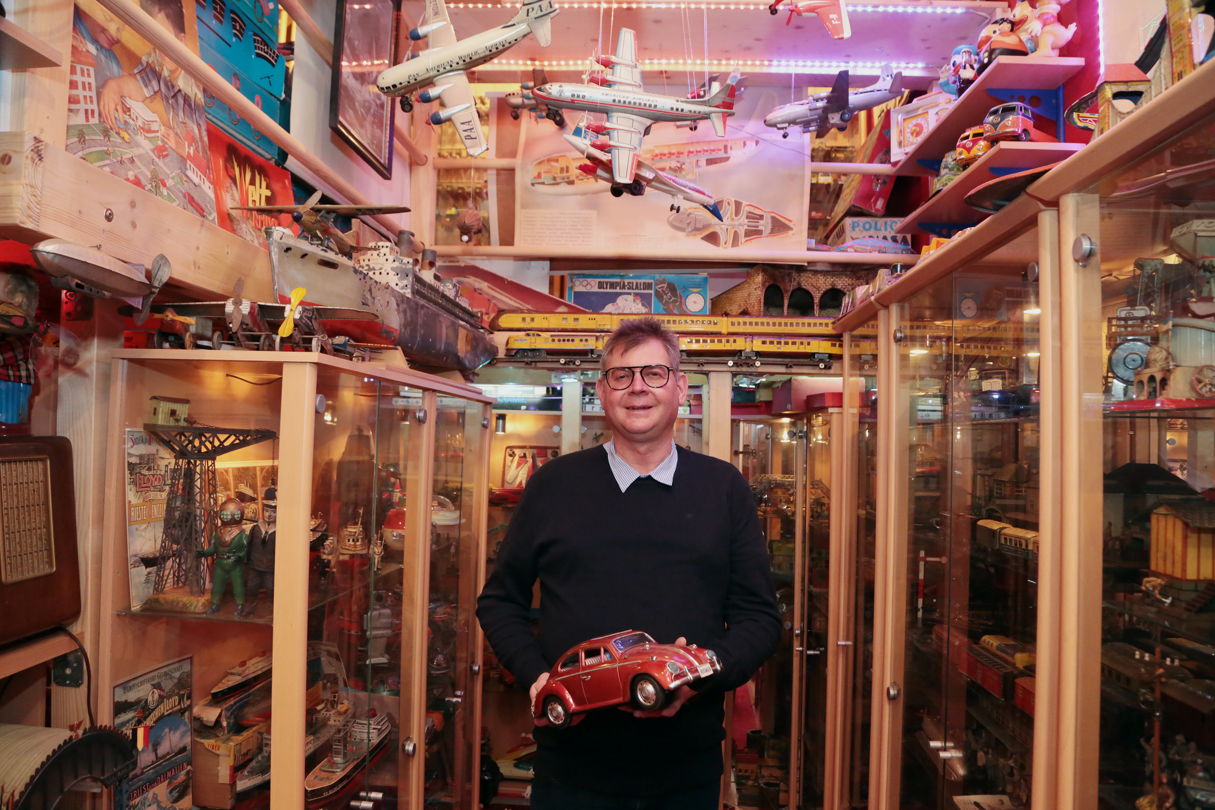 Johannes Krämmer steht mit einem Spielzeugauto in der Hand vor meterhohen Vitrinen mit unzähligen Spielzeugexponaten