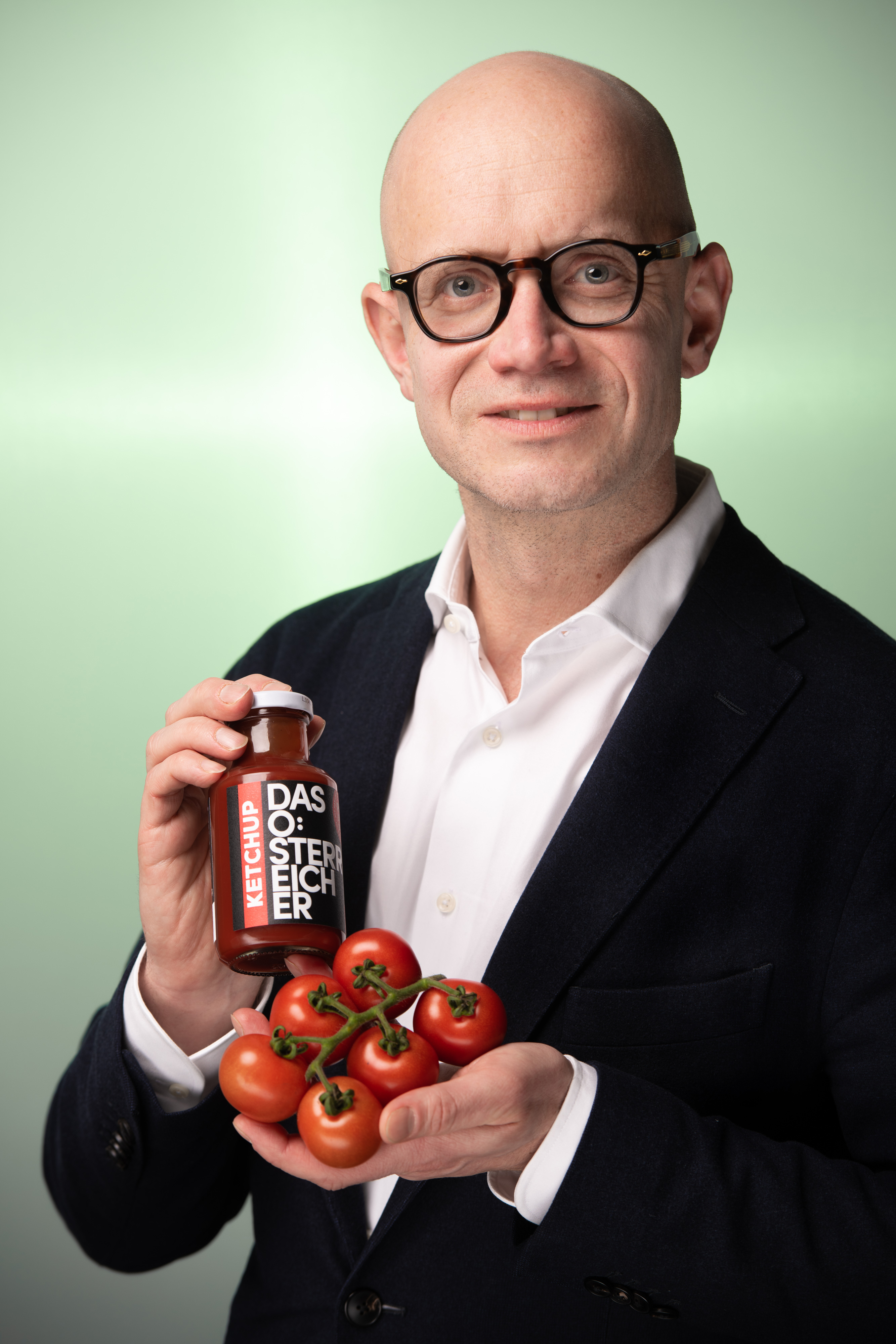 Peter Spak steht mit einem Glas Tomatensauce und einem Bündel Tomaten in der Hand vor einem grünen Hintergrund