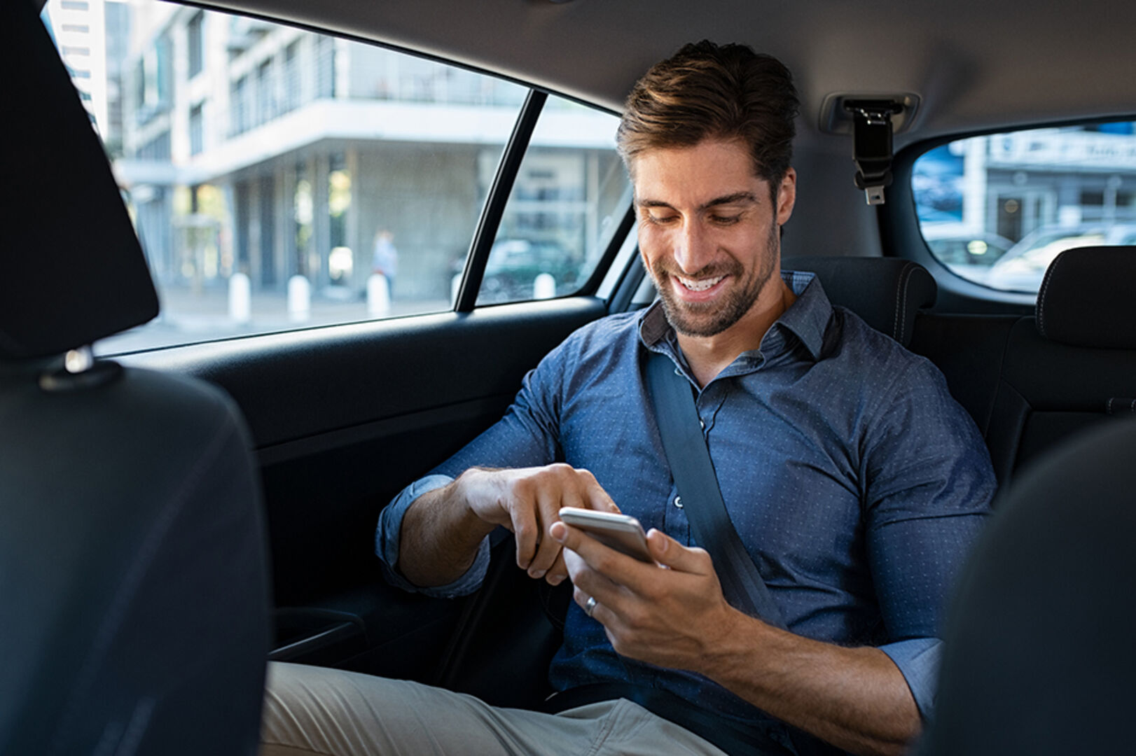 Mann mit Blick auf sein Smartphone auf dem Rücksitz eines Autos | Credit: iStock.com/Ridofranz