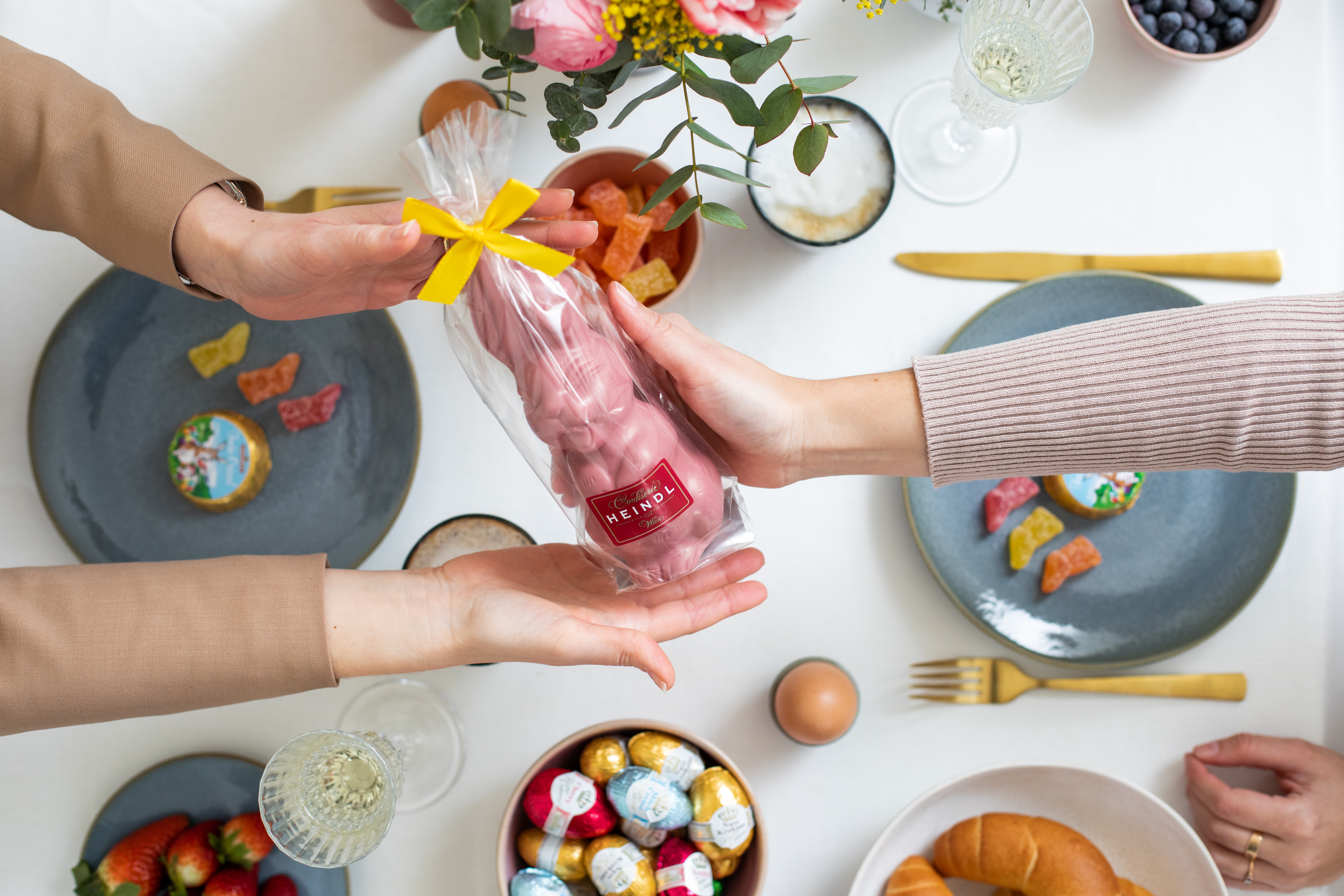 Eine Hand reicht zwei anderen einen rosaroten Schokohasen der Confiserie Heindl über einen festlich gedeckten Ostertisch