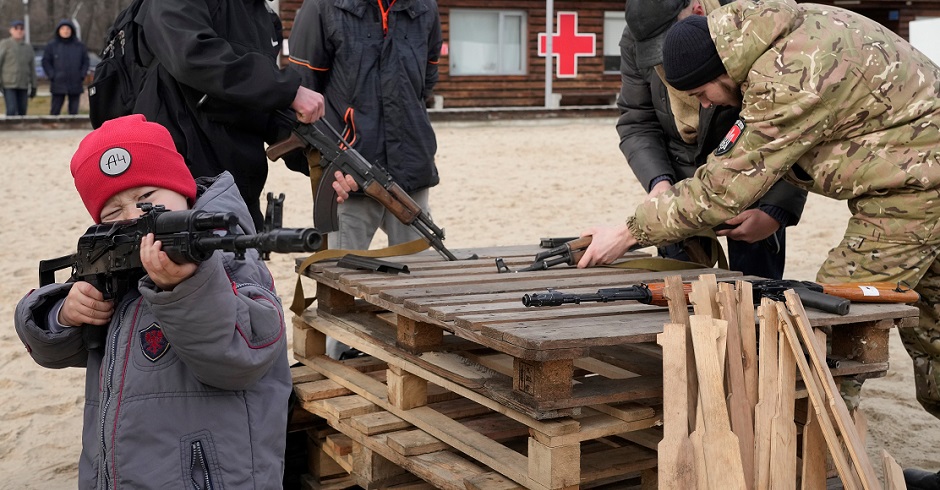 Ein Bub zielt mit einer Waffe in die Ferne, im Hintergrund beraten drei junge Männer und ein Soldat über die Lage.