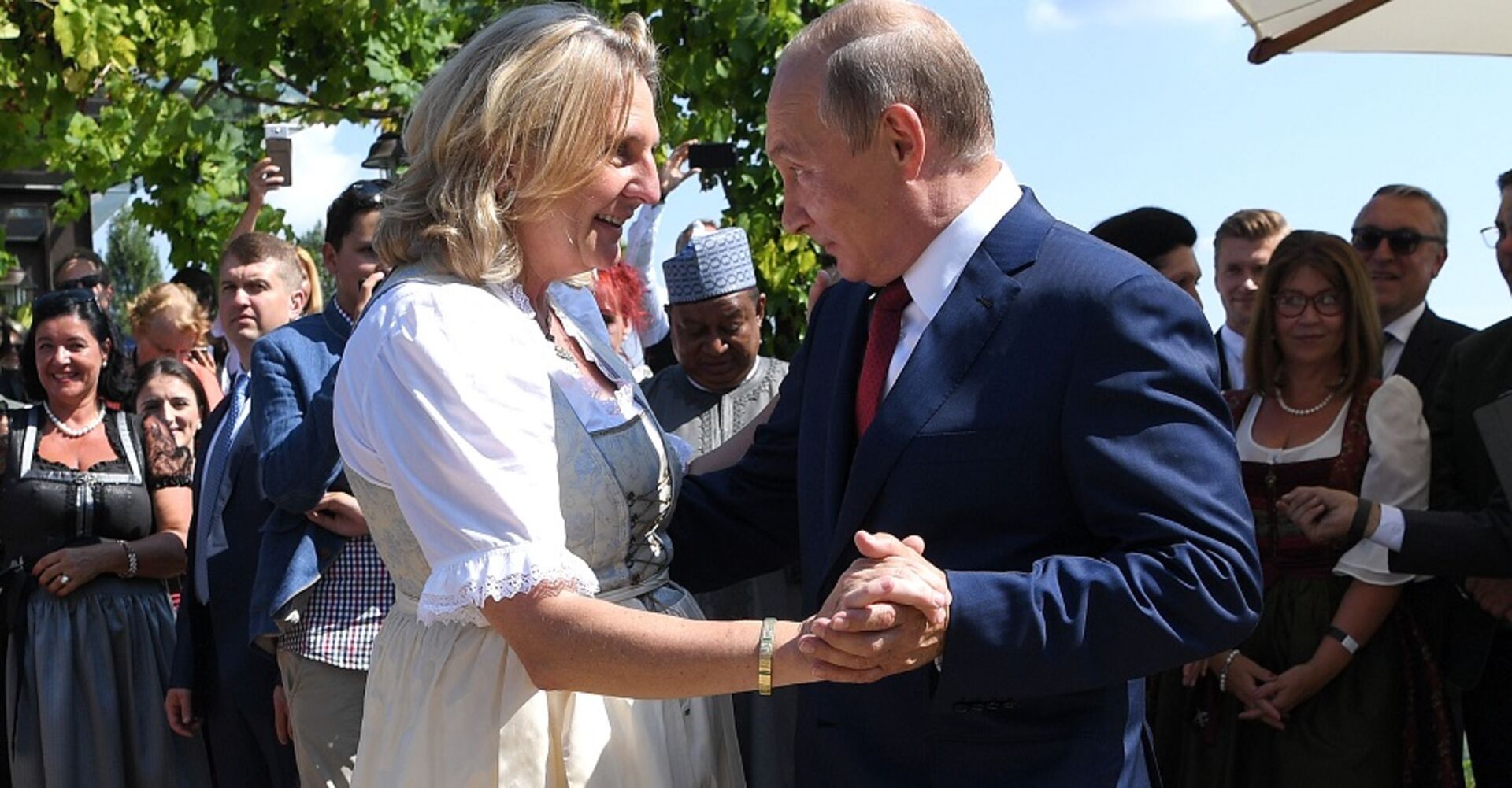 Karin Kneissl auf ihrer Hochzeit in einem Dirndl. Sie tanzt mit Wladimir Putin. Ringsum stehen Hochzeitsgäste und schauen zu.