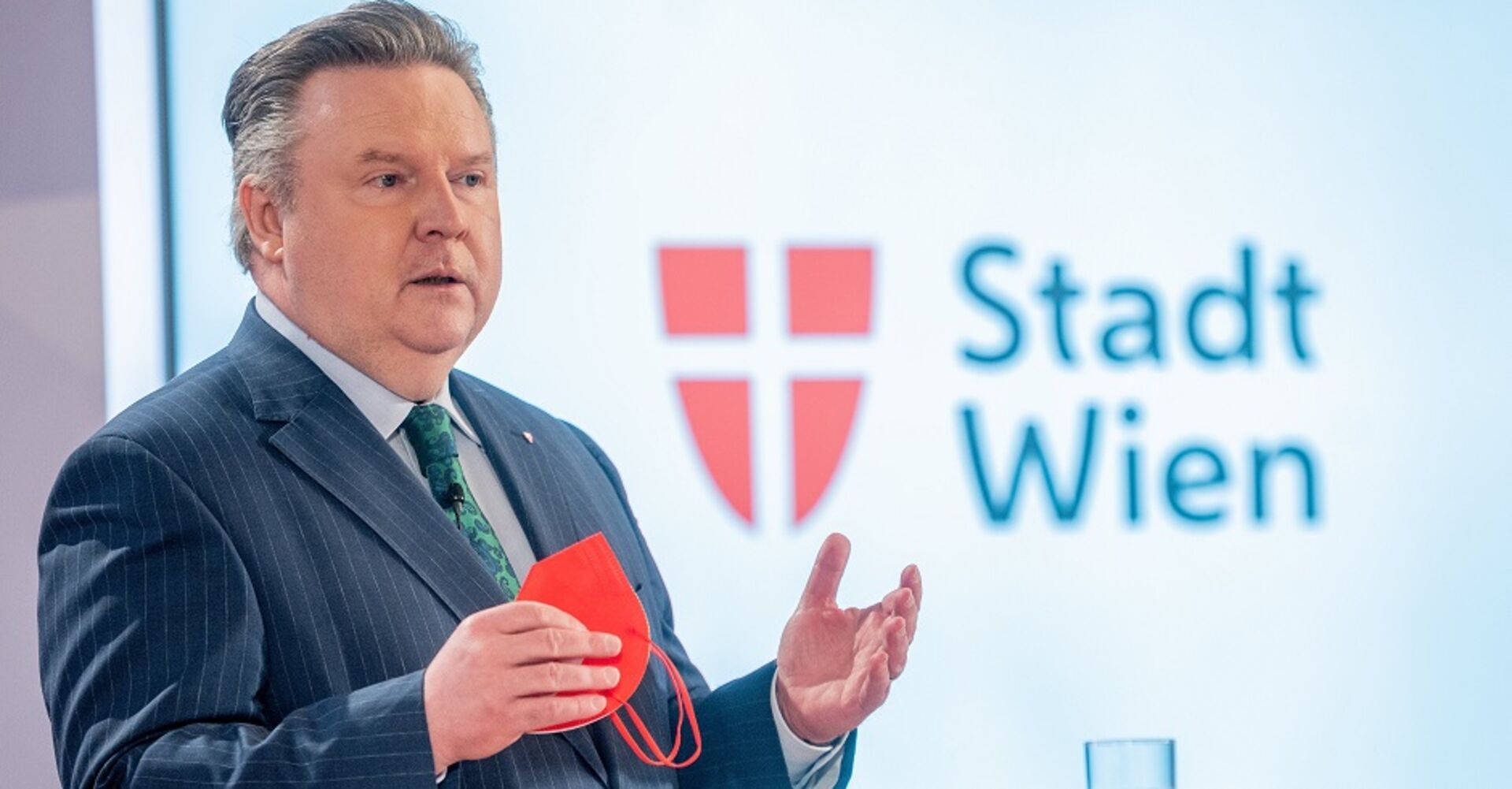 Bürgermeister Michael Ludwig erklärt auf einer Pressekonferenz die aktuelle Situation. Er hält eine rote FFP2-Maske in der Hand. Im Hintergrund der Schriftzug &quot;Stadt Wien&quot;, daneben das Wiener Stadtwappen.