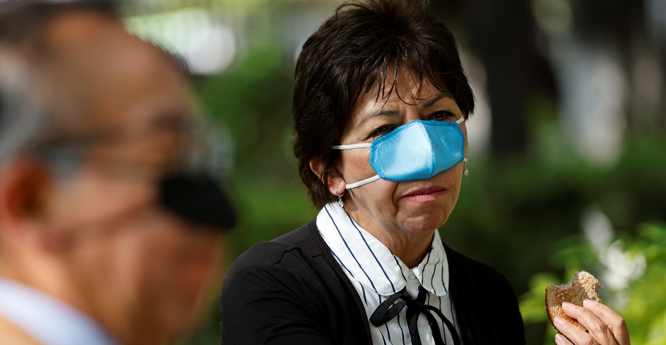 Eine asiatische Frau trägt eine hellblaue, untypisch kleine Maske über der Nase. Der Mund ist unbedeckt. In der Hand hält sie ein Stück Brot.