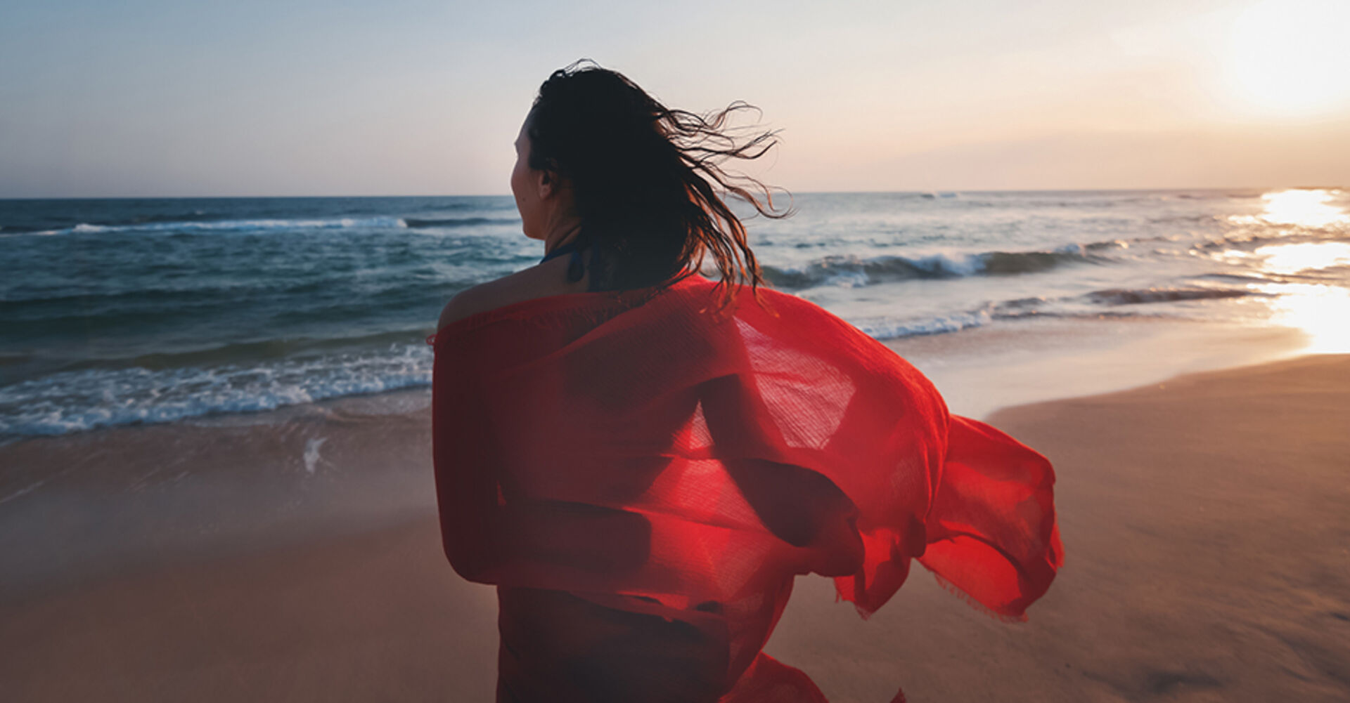 Junge Frau mit leichtem rotem Umhang spaziert bei Sonnenuntergang den Strand entlang | Credit: iStock.com/Olezzo