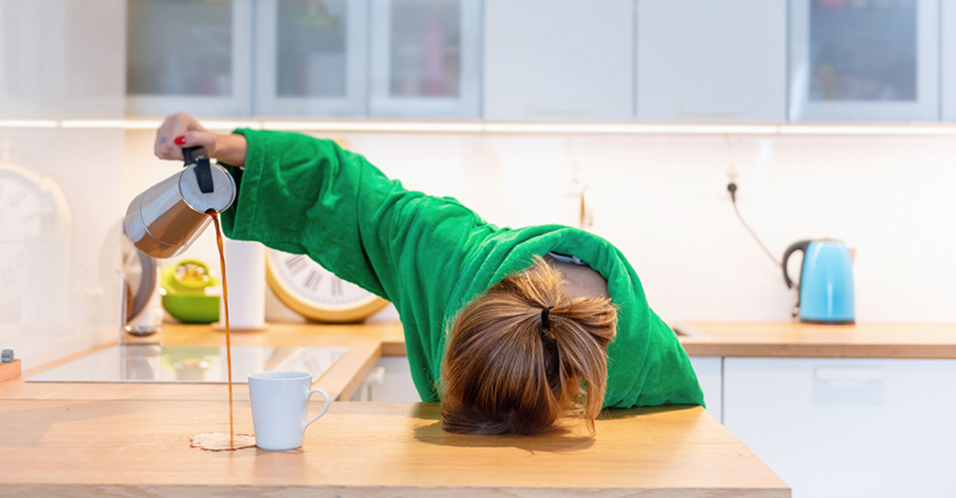 Müde Frau hat ihren Kopf auf die Tischplatte gelegt und verschüttet beim Einschenken den Kaffee | Credit: iStock.com/morgan23