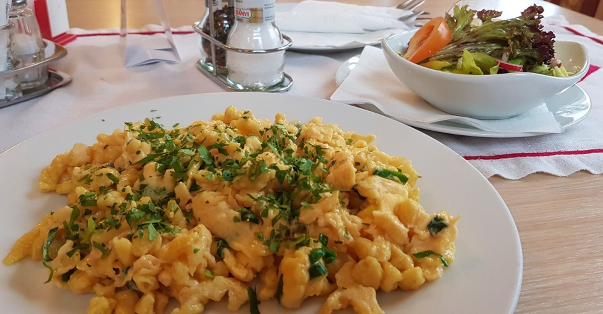 Ein Teller Eiernockerl mit Schnittlauch-Garnitur auf einem Wirtshaustisch. Dazu gibt es gemischten Salat. Griffbereit: Salz und Pfeffermühle.