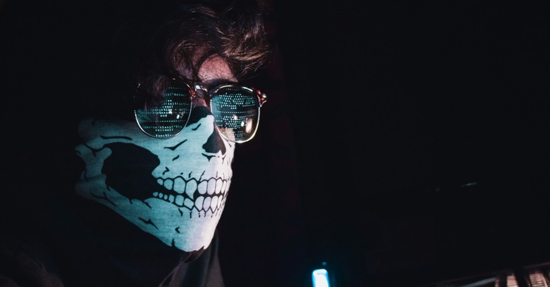 Ein junger Mann mit einer Totenkopfmaske und einer Brille. In den Gläsern spiegelen sich Zahlen und der Code auf einem Computerbildschirm