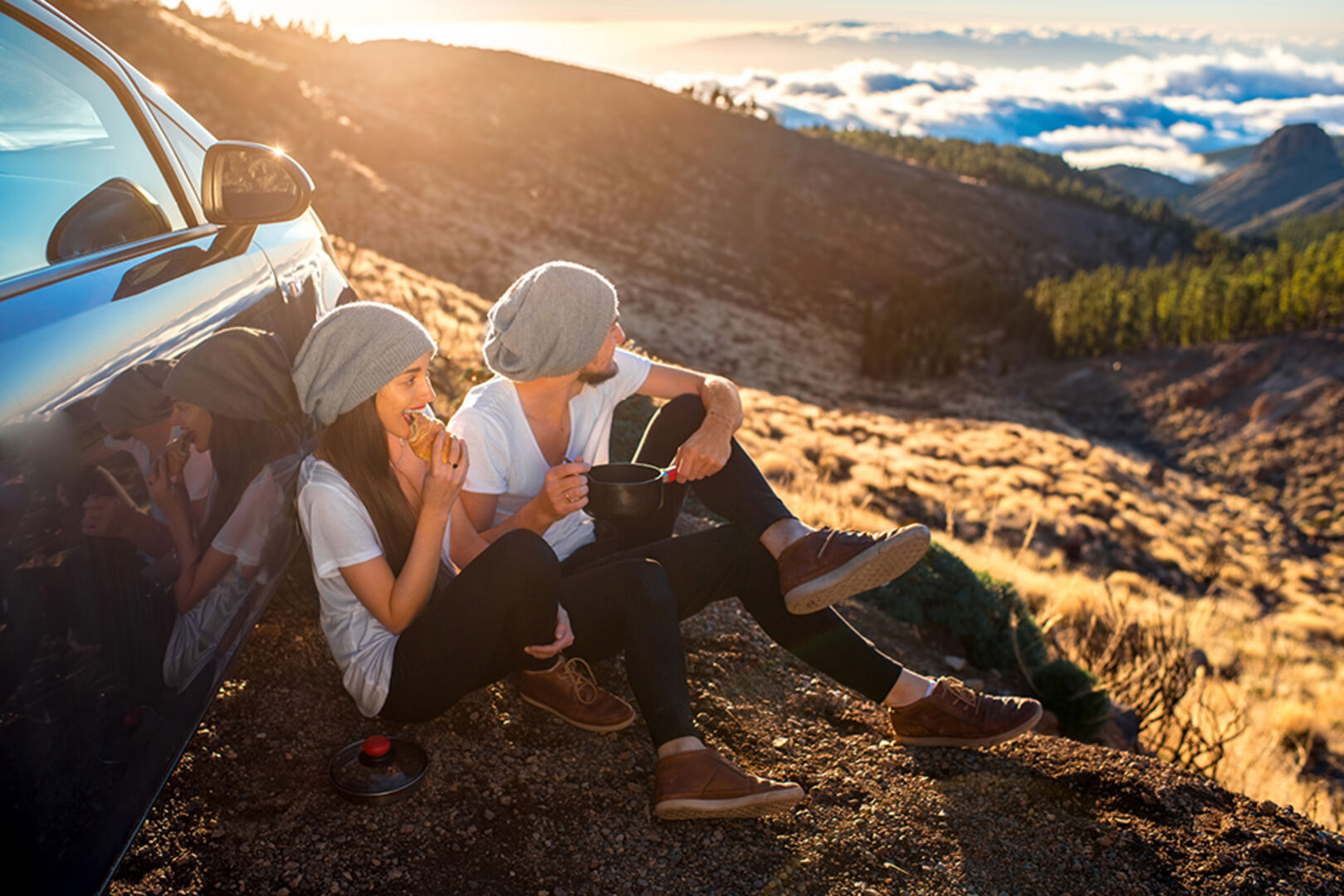 Junges Pärchen sitzt vor seinem Auto mit Bergpanorama | Credit: iStock.com/RossHelen