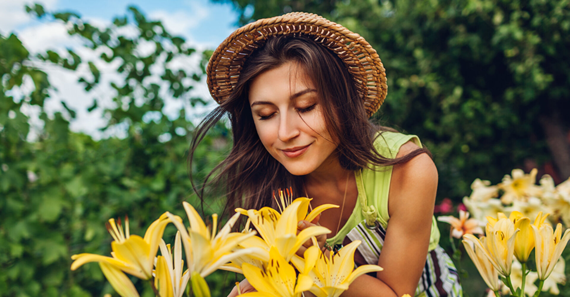 Junge Frau mit Sommerhut riecht an den blühenden Lilien in ihrem Garten | Credit: iStock.com/Maryviolet