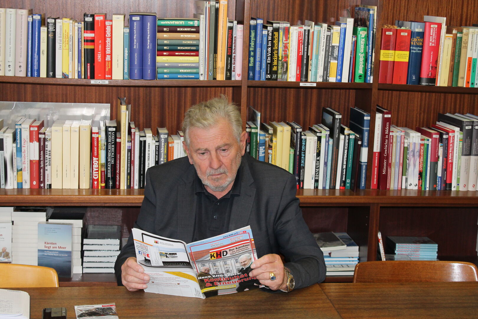 Andreas Mölzer sitzt an einem Tisch vor einem Bücherregal und blättert in der Mitgliederzeitung des KHD