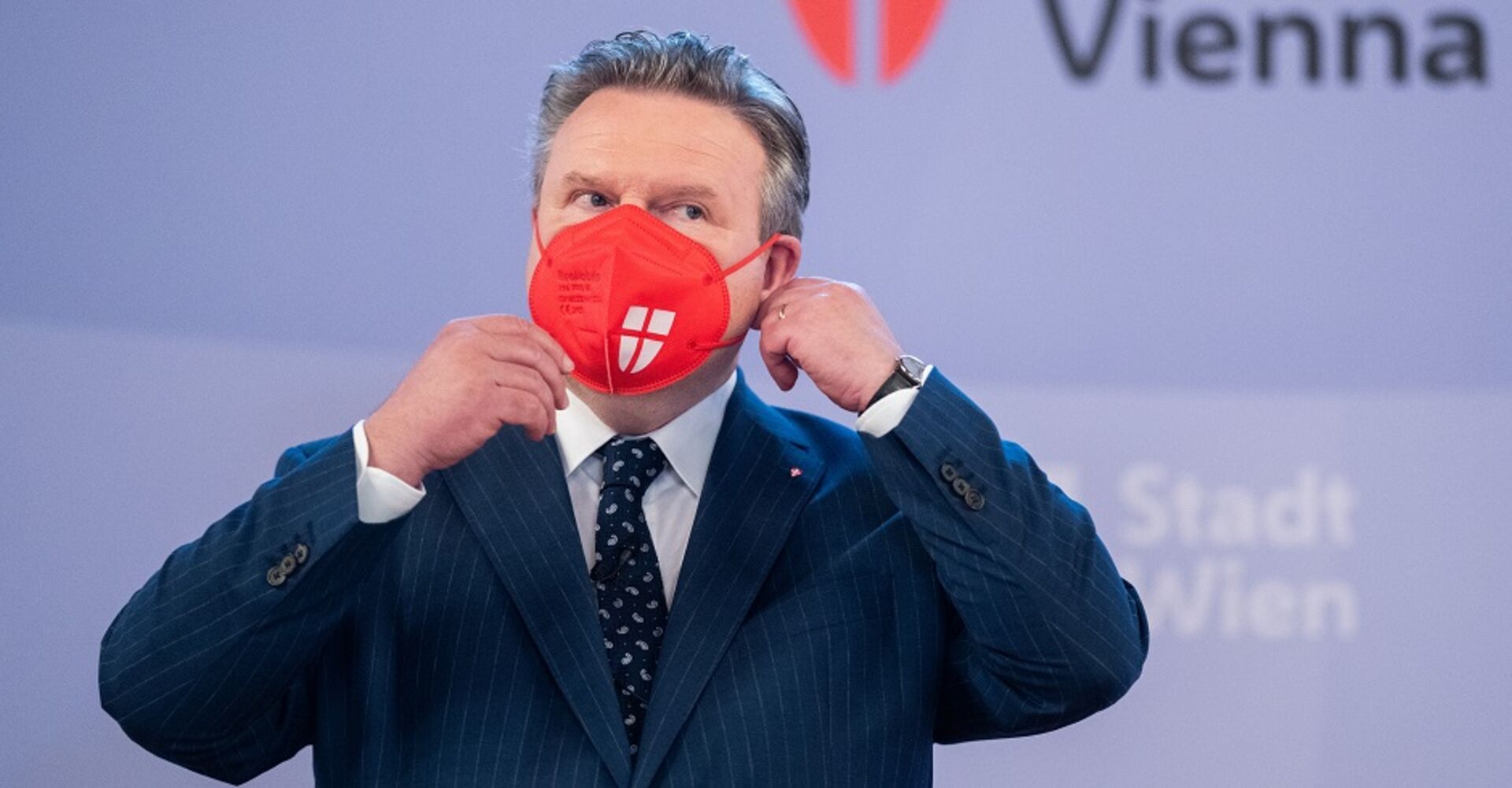 Michael Ludwig nimmt eine rote FFP2-Maske vor der Pressewand der Stadt Wien ab.