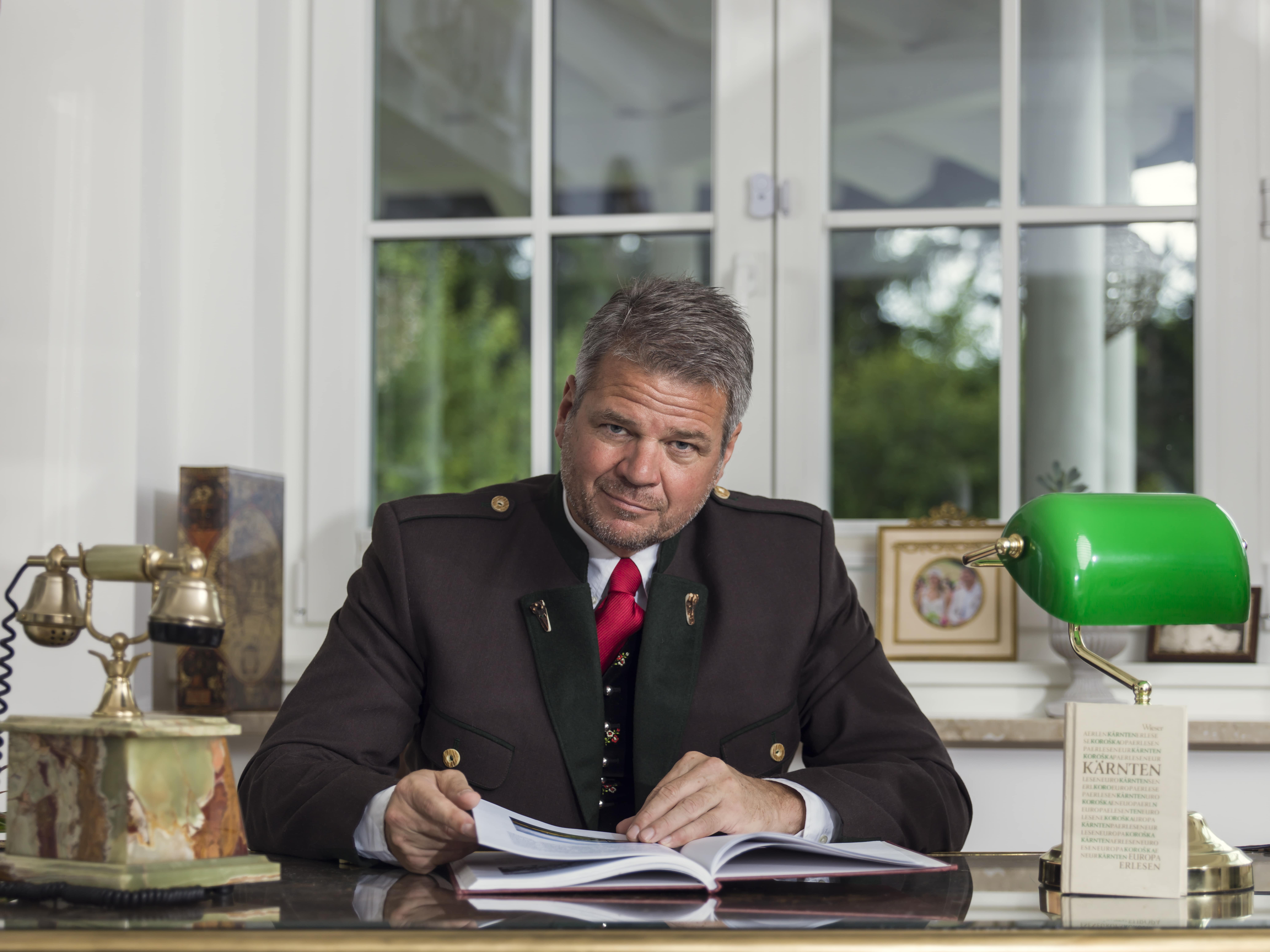 Bürgermeister Köfer sitzt in einem Kärntner Anzug hinter seinem Schreibtisch und blättert in einem Buch