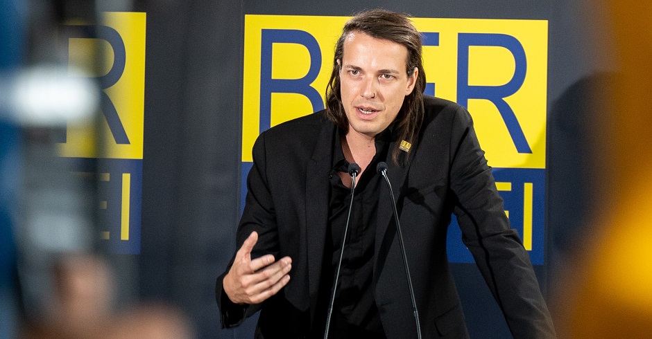 Bierpartei Gründer Marco vor Kameras bei der Verkündung seiner Kandidatur. Er trägt ein schwarzes Hemd und schwarzen Anzug.