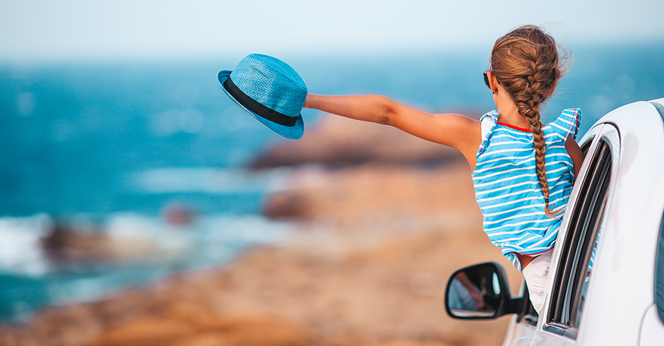 Kleines Mädchen lehnt sich entspannt mit ihrem Sommerhut aus dem Auto und blickt Richtung Meer | Credit: iStock.com/travnikovstudio