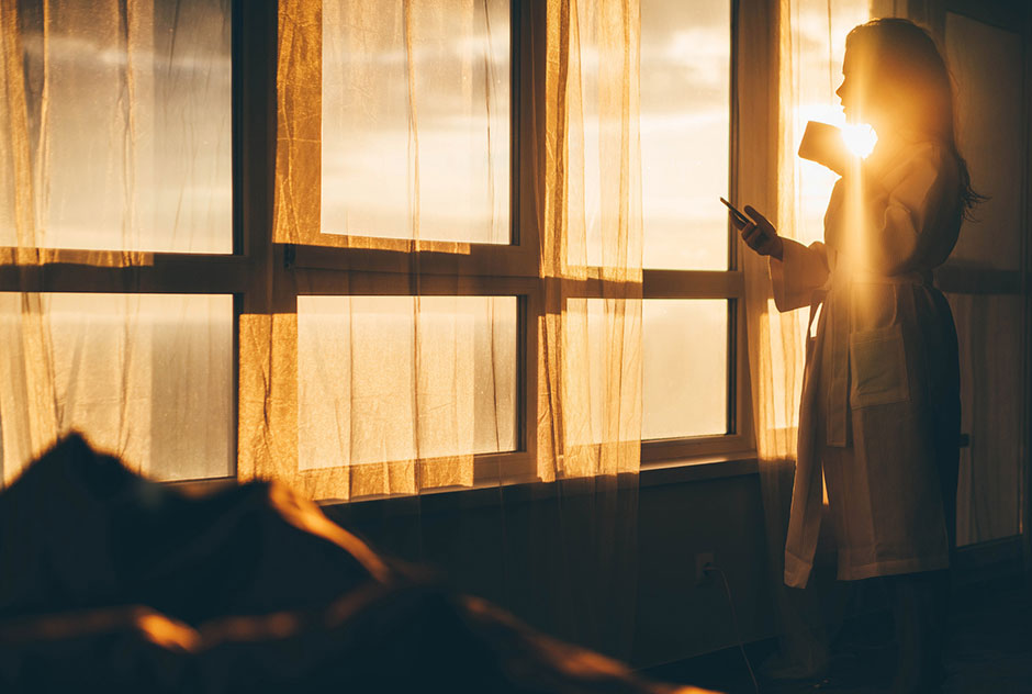 Mit einer Tasse Kaffee in der Hand genießt eine junge Frau den Sonnenaufgang vom Fenster ihres Schlafzimmers aus | Credit: iStock.com/Maria Korneeva