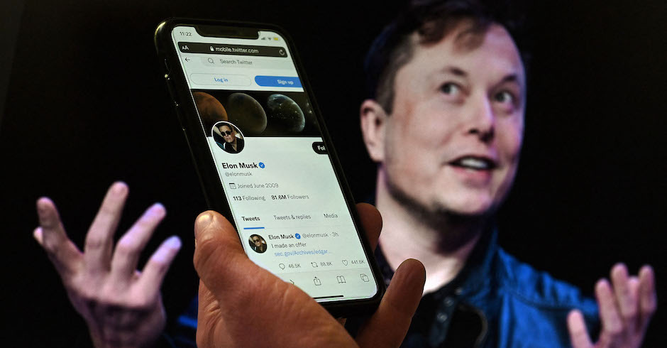 Eine Hand hält ein Smartphone auf dem Elon Musks Twitter-Account zu sehen ist. Im Hintergrund Elon Musk