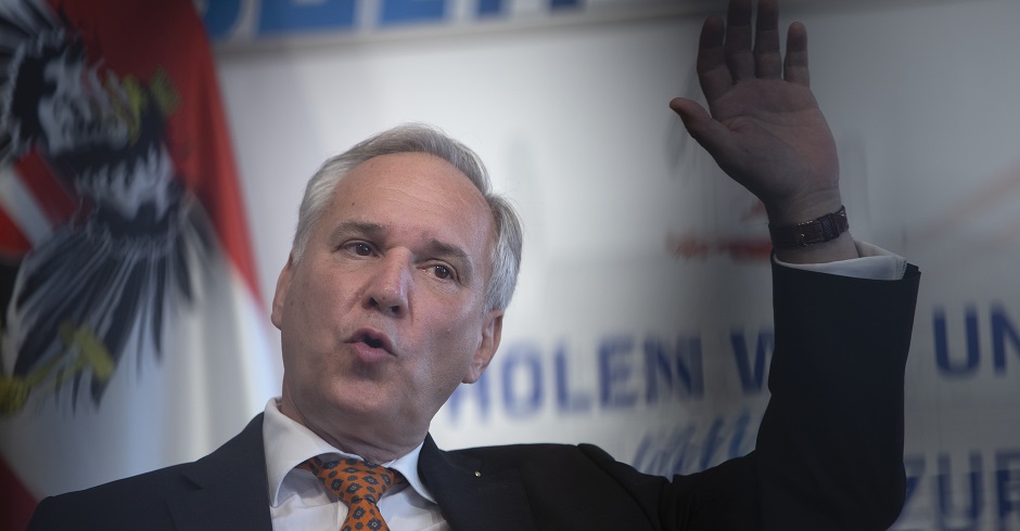 Walter Rosenkranz vor der Pressewand des Wahlkampfs. Im Hintergrund die österreichische Flagge. Er spricht und hat die linke Hand in der Luft erhoben.