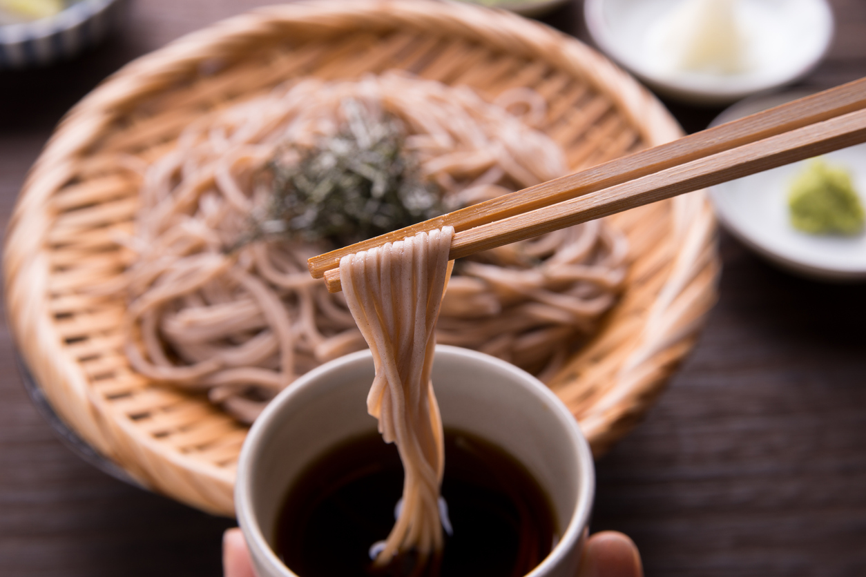 Ein Korb mit japanischen Soba Nudeln sowie zwei Essstäbchen die Nudeln in eine Suppenschale eintauchen