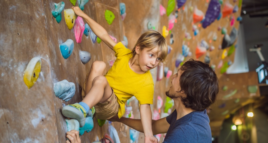 Vater hilft Sohn in der Kletterhalle beim Klettern. | Credit: iStock.com/galitskaya