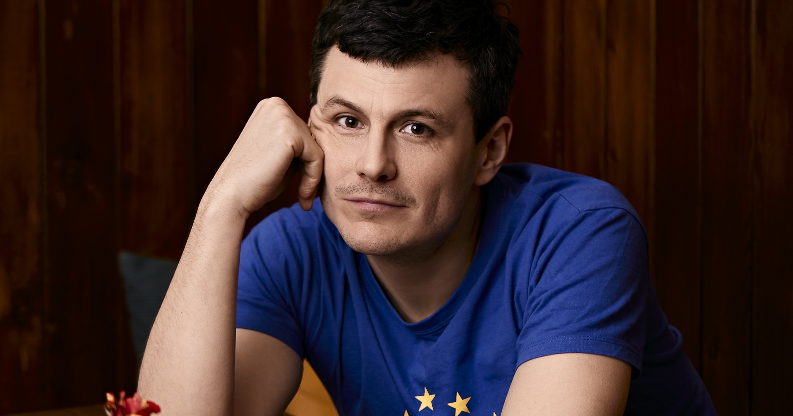 Manuel Rubey sitzt an einem Tisch, stützt den Kopf auf die Hand und schaut in die Kamera