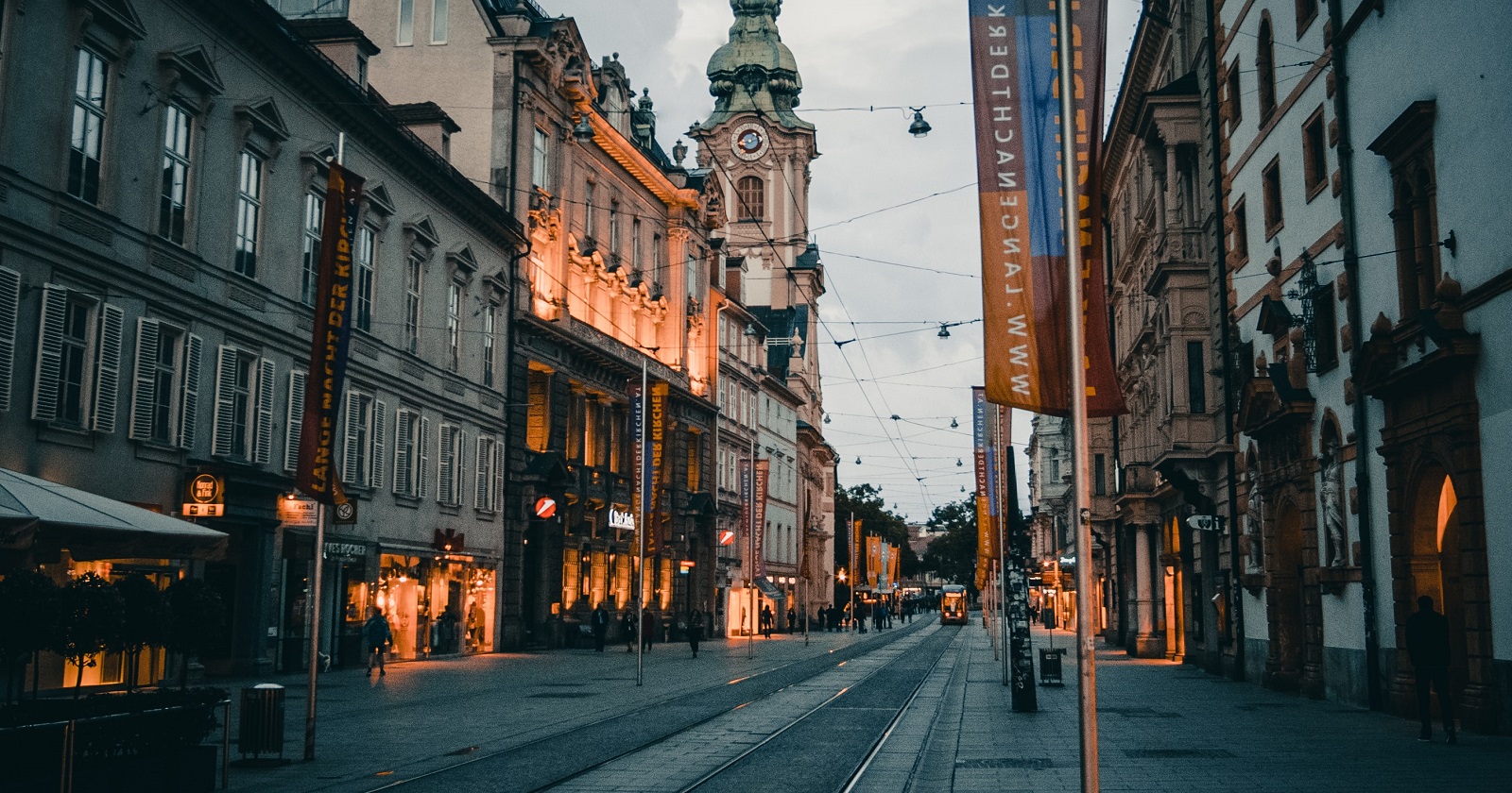 Blick in die Grazer Herrengasse bei Dämmerung. Einzelne Geschäfte sind beleuchtet, die Straßenbeleuchtung ist dunkel