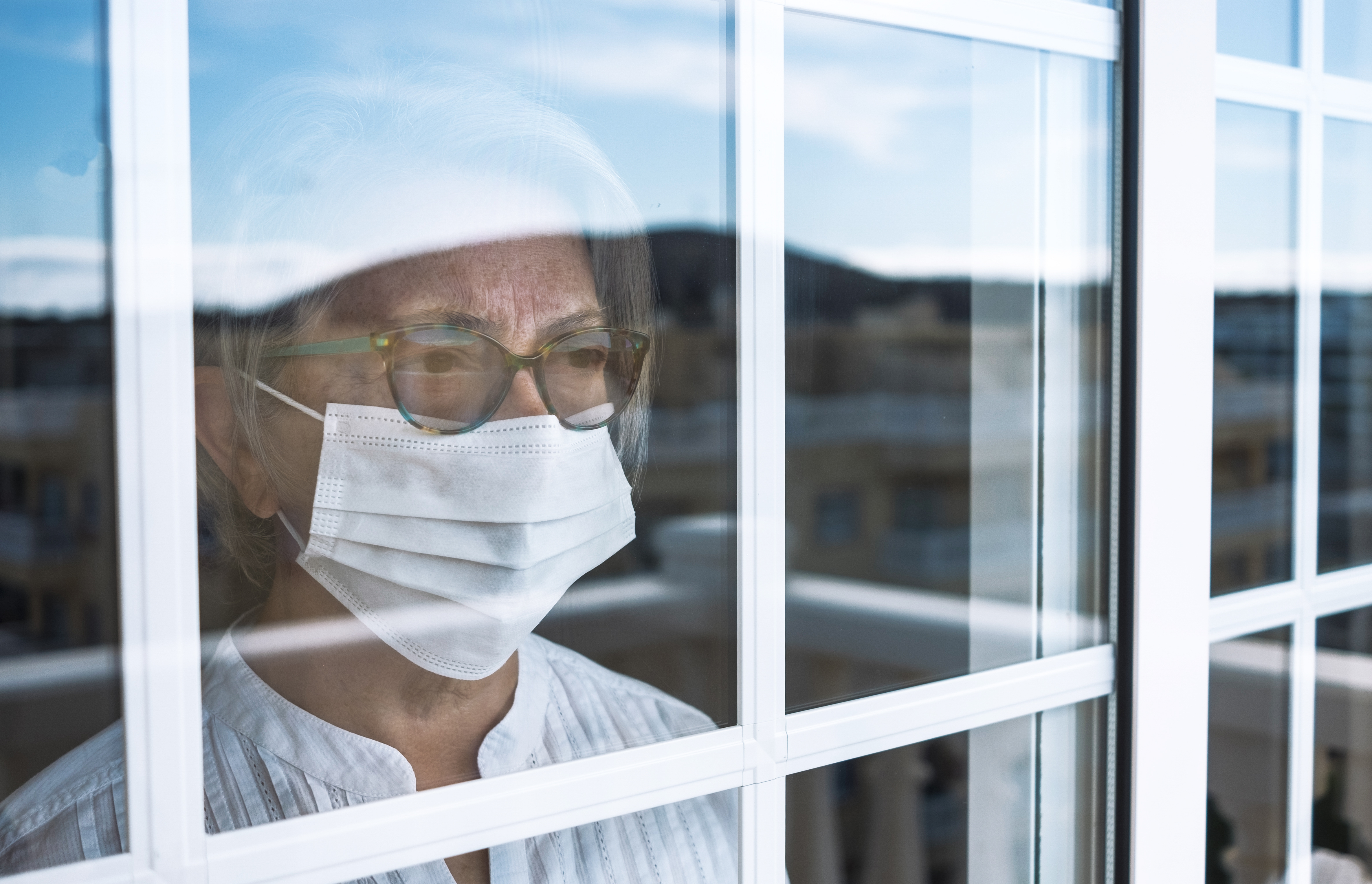 Ältere Frau mit Mundschutz-Maske blickt aus dem Fenster. | Credit: iStock.com/lucigerma