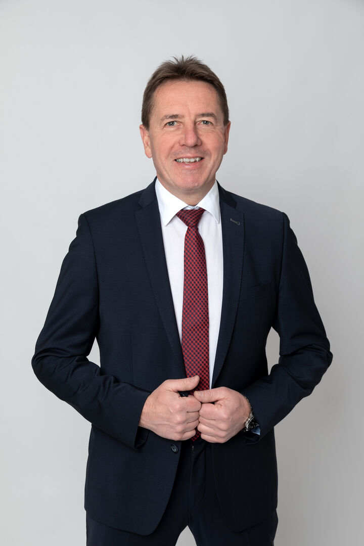 Erwin Angerer, der einen schwarzen Anzug mit weißem Hemd sowie roter Krawatte trägt und vor einem grauen Hintergrund steht