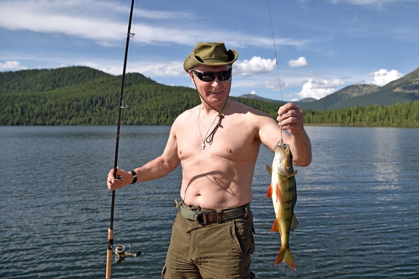 Putin mit Hut, Sonnenbrille und Angel. Er hat den Oberkörper entblößt und hält einen Fisch in der Hand.