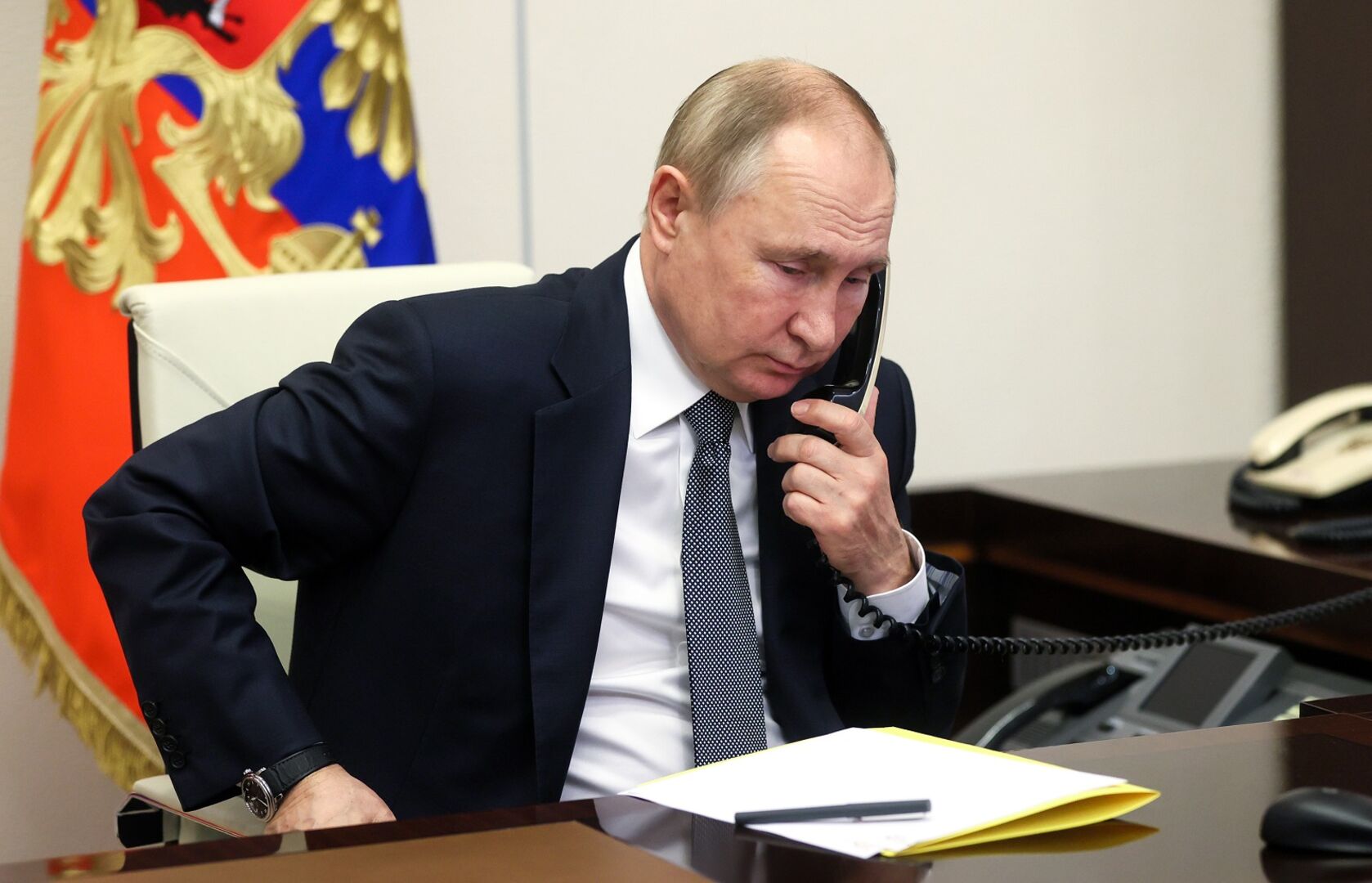 Putin sitzt an einem Schreibtisch und telefoniert mit einem Schnurtelefon.