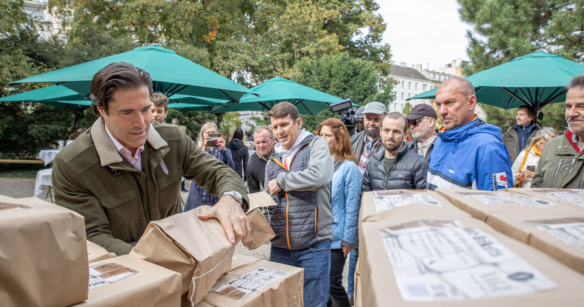 Tassilo Wallentin vor einem Stapel mit braunen Paketen. Er greift nach einem. Im Hintergrund warten Menschen.